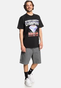 Black T-shirt with graphic print in purple, red, and white, paired with gray denim shorts. Model wears white sneakers and black socks.