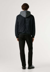 Chaqueta azul marino con capucha gris, puños y dobladillo elásticos, combinada con pantalones negros. El diseño presenta un exterior liso y un ajuste relajado.