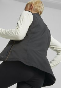 Czarna kamizelka bez rękawów z wysokim kołnierzem, wyposażona w kieszenie na zamek i zaokrąglony dolny brzeg, noszona na jasnej koszuli z długim rękawem.