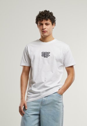 Balta medvilninė marškinėliai trumpomis rankovėmis ir apvaliu kaklu, su juodu išsiuvinėtu „nyc“ logotipu centre. Dėvimi su šviesiai mėlynais džinsais.