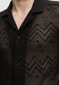 Gros plan d'une personne portant une chemise noire en crochet boutonnée avec des motifs en zigzag et un col, montrant la peau nue en dessous.