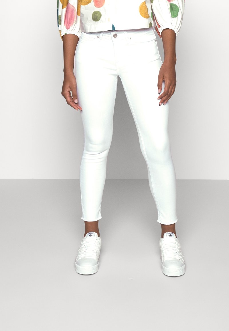 Los vaqueros blancos ajustados tienen un corte slim con un dobladillo recortado, combinados con zapatillas blancas. El modelo lleva una blusa colorida de lunares.