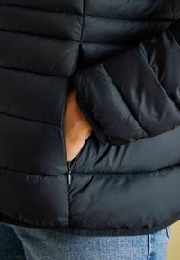 Juodas pūkuotas švarkas su horizontaliu susiuvimo raštu, šoniniu užtrauktuku ir minkštu pamušalu ant rankogalių. Detali medžiagos nuotrauka.