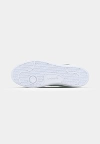 Semelle d'une chaussure Lacoste blanche avec motif à quadrillage et détail circulaire près du talon sur un fond gris clair.