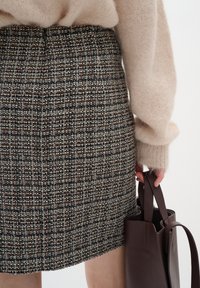 Knee-length tweed seelik tumeses pruunis, kreemjas ja punases mustris; pehme beežs kampsun puffsete varrukatega; hoiab pruuni nahkkotti.