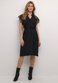 Schwarzes Kleid mit Spitzenkurzärmeln, V-Ausschnitt und einem Bindegürtel in der Taille. Der Stoff hat eine dezente Textur und reicht bis zur Wadenmitte. Kombiniert mit schwarzen Knöchelstiefeln.