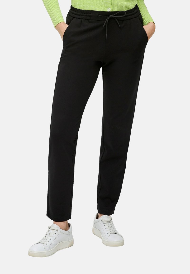 Pantalons de jogging noirs, fuselés, avec une taille élastique et un cordon de serrage. Matière lisse, poches latérales, associés à des baskets blanches.