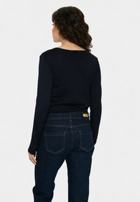 Tamsiai mėlyno ilgasleeve megztinis su bangų formos apykakle, dėvimas su tamsiai mėlynomis džinsinėmis kelnėmis su subtiliu siuvinėjimu ir dviem užpakalinėmis kišenėmis.