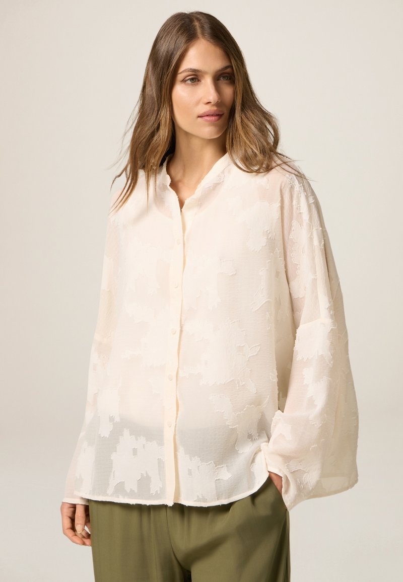 Blusa leggera color pesco con una texture a motivo floreale, apertura con bottoni sul davanti e maniche lunghe, realizzata in un tessuto morbido e trasparente con una vestibilità rilassata.