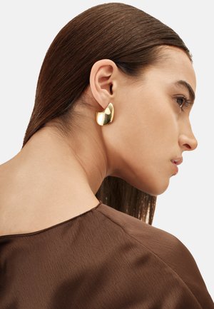Pendientes de tono dorado con una forma curva y suave, que exhiben un acabado brillante. Estilizados en una modelo con cabello liso y oscuro. Se ve una blusa de tela marrón.