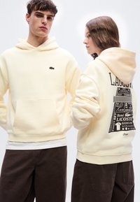 Νέος άνδρας και γυναίκα που φορούν κρεμ φούτερ με κουκούλα και σκούρα παντελόνια, με το λογότυπο Lacoste στο εμπρόσθιο μέρος και μαύρο γραφικό με θέμα το τένις στην πλάτη.
