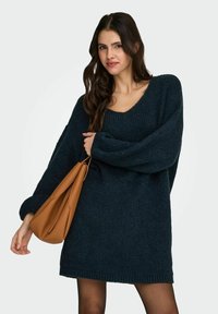 Granatowy, oversize'owy sweter z dzianiny z dekoltem w serek, długimi rękawami i prążkowanym wykończeniem na dole. Do tego jasnobrązowa skórzana torebka.