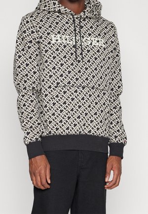 Schwarz-weiß gemusterter Kapuzenpullover mit "TOMMY HILFIGER"-Logo auf der Brust, getragen zu schwarzen Hosen, Hände locker an den Seiten.