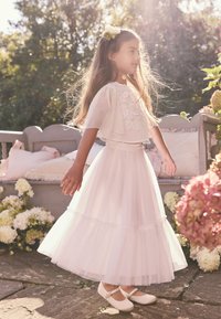Falda de tul blanca en capas con top corto bordado de flores, combinada con zapatos Mary Jane blancos. Entorno natural con acentos florales.