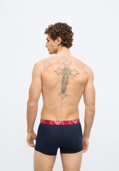 Cuecas boxer azul-marinho com uma cintura vermelha que apresenta detalhes do logotipo. O modelo tem cabelo encaracolado e tatuagens visíveis nas costas.