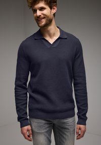 Mörkblå, långärmad stickad tröja med V-ringning och krage. Texturerat tyg. Bärs med ljusgrå jeans, poserad mot en neutral bakgrund.