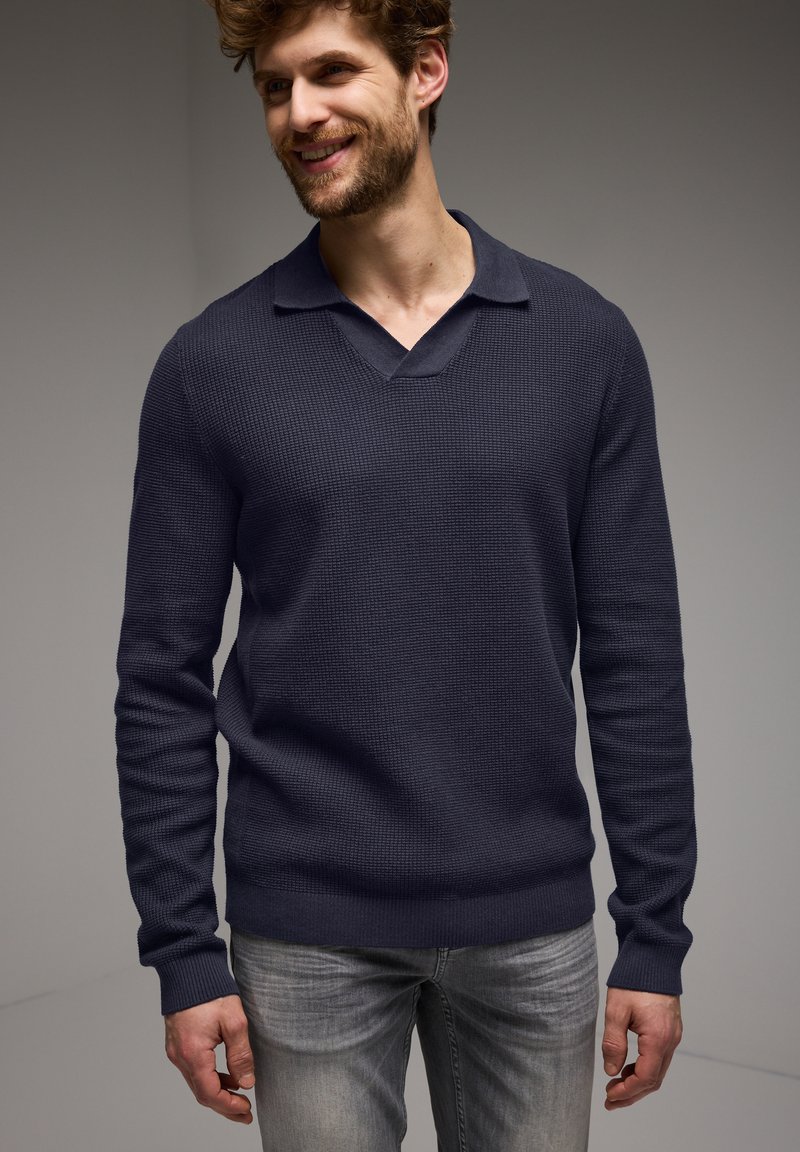 Mörkblå, långärmad stickad tröja med V-ringning och krage. Texturerat tyg. Bärs med ljusgrå jeans, poserad mot en neutral bakgrund.