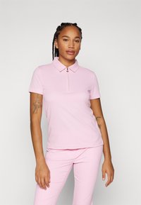 Camisa polo rosa claro de mangas curtas com colarinho com zíper, apresentando um tecido texturizado. Usada com calças rosa claro combinando.
