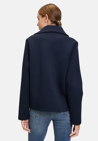 Dunkelblaue, cropped Jacke mit einem breiten Kragen und langen Ärmeln, aus weichem Stoff. Getragen über hellblauen Jeans.