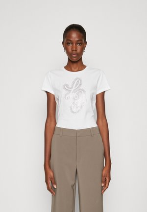 T-shirt bianca a maniche corte con un design grafico argento che presenta "LJ" in un font decorativo, abbinata a pantaloni beige.