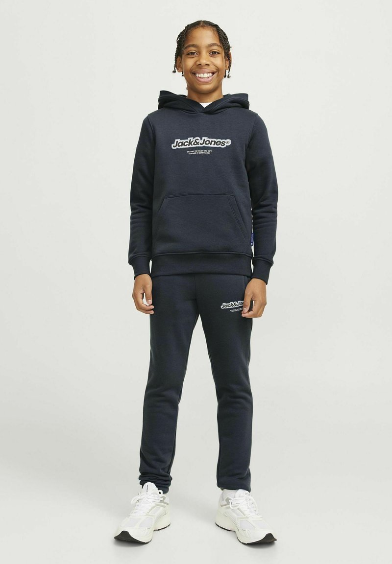 Marineblå hættetrøje med en forlommer, hvidt logo-tekst, matchende sweatpants og hvide sportsko med mørke accenter.