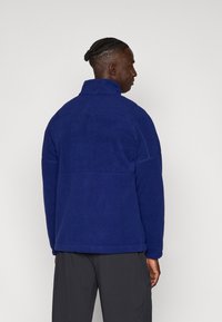 Chaqueta de forro polar azul marino con cuello alto, corte holgado y costuras mínimas, con mangas largas y una textura lisa.