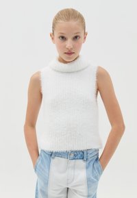 Top blanco sin mangas con un alto cuello de pelaje; textura acanalada y bordes deshilachados; combinado con pantalones de denim de estilo patchwork en azul claro.