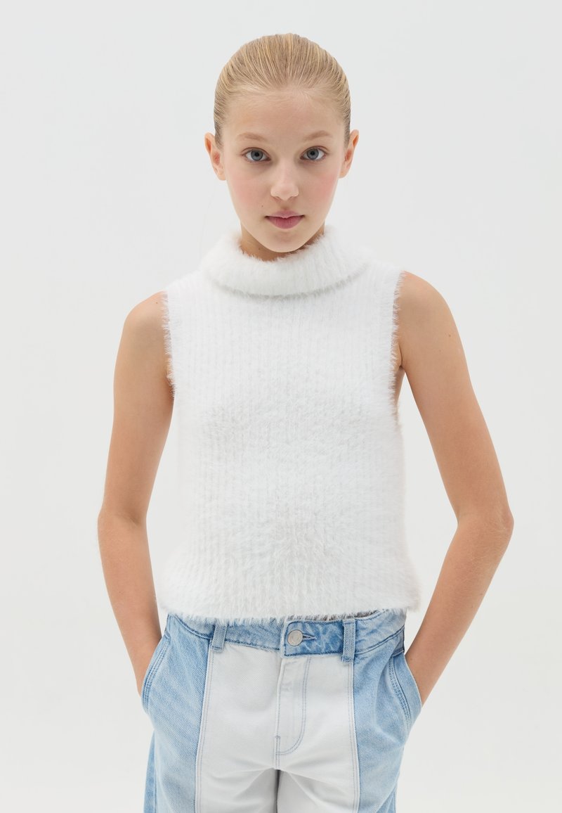 Top blanco sin mangas con un alto cuello de pelaje; textura acanalada y bordes deshilachados; combinado con pantalones de denim de estilo patchwork en azul claro.