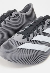 Chaussures de sport grises avec texture côtelée, lacets noirs et motif blanc à trois bandes, montrées en gros plan.
