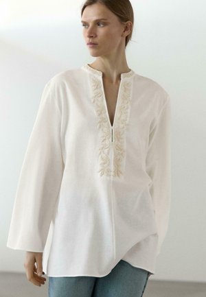 Woman wearing a loose, long-sleeve white blouse with floral embroidery around the V-neck, paired with light blue jeans.