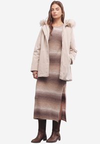 Parka impermeabile beige con cappuccio bordato di pelo, indossata sopra un vestito lungo con gradazione marrone e spacco laterale, abbinata a stivali marroni al ginocchio.