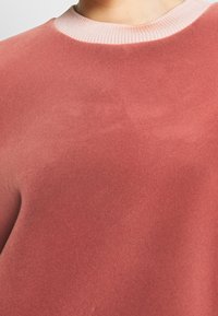 Sudadera rosa con textura suave, que presenta un cuello acanalado en un tono más claro. El diseño es sencillo y casual, sin patrones ni adornos.