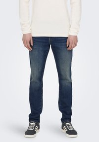 Dunkelblaue Jeans im Slim-Fit mit leichten Ausbleichungen an den Oberschenkeln, kombiniert mit einem cremefarbenen, strukturierten Langarmhemd und grauen Turnschuhen.
