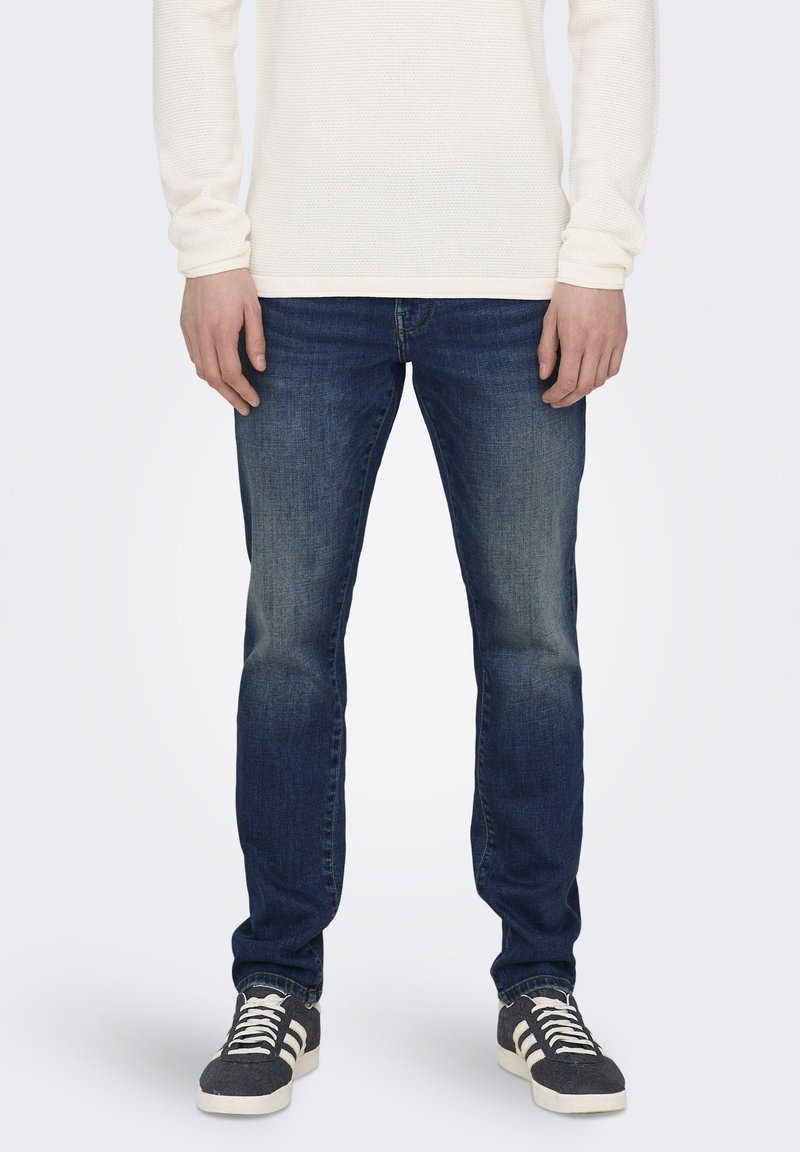 Dunkelblaue Jeans im Slim-Fit mit leichten Ausbleichungen an den Oberschenkeln, kombiniert mit einem cremefarbenen, strukturierten Langarmhemd und grauen Turnschuhen.