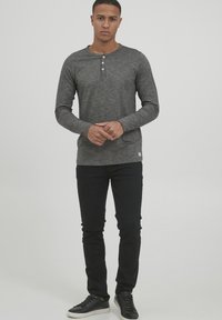 Langærmet, grå henley skjorte med rund halsudskæring og tre-knaps placket, parret med sorte jeans og sorte sneakers.