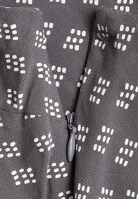 Harmaa kangas, jossa on geometrinen valkoinen kuvio, varustettuna vetoketjulla ja tekstuuripinnalla. Suunnittelussa on erilaisia pisteitä ja muotoja.