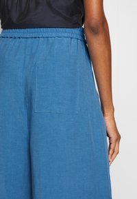 Blauwe linnen broek heeft een elastische tailleband, een achterzak en een relaxte pasvorm, met een gladde textuur.