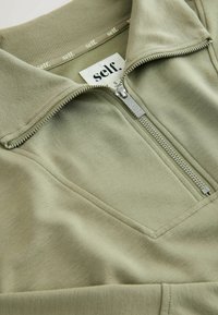 Olivově zelený pulovr na zip s žebrovaným límcem, hladkým povrchem a viditelným půlzipem. Obsahuje značkovou etiketu uvnitř límce.