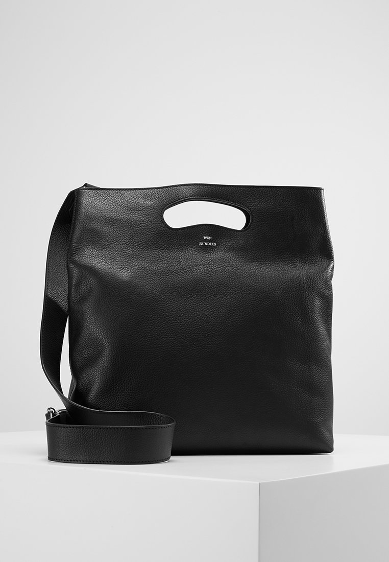 Sac cabas en cuir noir avec poignée intégrée et bandoulière détachable, présenté sur une surface blanche avec un fond gris.
