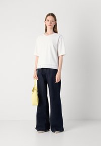 Hvit kortermet topp med detaljert nederkant, kombinert med mørkeblå vide jeans. Holder en gul håndveske med et stilrent design.