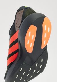 Sports shoe with a black upper, orange stripes, and a textured rubber sole featuring orange and white accents. The design includes perforations.