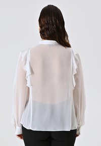 Una donna con i capelli bagnati indossa una blusa bianca trasparente con spalle arricciate e maniche lunghe, abbinata a pantaloni neri, voltata di spalle.