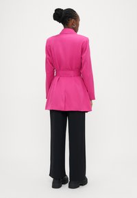 Ροζ σακάκι με ζώνη και εφαρμοστή γραμμή, με γιακά με εγκοπή, μακριά μανίκια και σκίσιμο στο πίσω μέρος, συνδυασμένο με μαύρο παντελόνι φαρδιάς γραμμής.