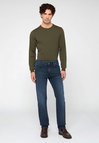 Camisa de manga larga verde oliva combinada con jeans rectos azul oscuro y zapatos de cuero marrón, en un fondo gris liso.