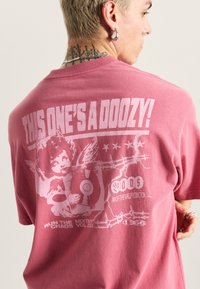 Lyserød bomulds-T-shirt med en stor hvid grafik med tekst og en illustration af en engel, der holder en plade. Løs pasform med rund hals.