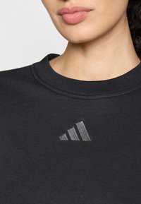 Felpa nera con scollatura tonda. Presenta un logo trefoil grigio texturizzato sul petto. Il tessuto appare morbido ed elastico.