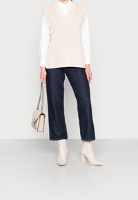 Personne portant un gilet en maille crème par-dessus une chemise blanche, un jean bleu foncé raccourci, des bottines à talons crème, tenant un sac à main beige avec une bandoulière chaîne.