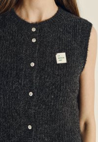 Donkergrijs gebreid vest met een ronde hals, knoopsluiting aan de voorkant en een klein groen-wit label waarop "LA MAISON AMV" staat.