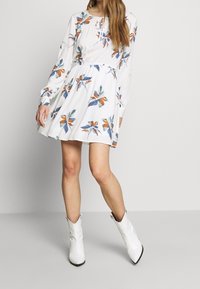 Mujer vestida con un vestido largo de manga larga blanco con estampados florales en naranja y azul, complementado con botas de tobillo blancas de punta afilada.