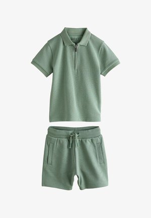 Polo shirt verde a maniche corte con zipper e pantaloni corti coordinati con coulisse e tasche laterali, posati su uno sfondo bianco.